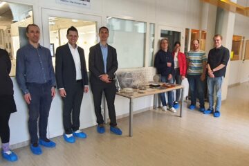 Prof. Dr. Brem (Executive Board IMS CHIPS & Vice-Rector for Transfer and International Affairs as well as Managing Director at ENI) and Prof. Dr. Nils Quack (Managing Director IMS CHIPS) are in the center of the picture, surrounded by employees of IMS CHIPS and colleagues from the Institute of Entrepreneurship and Innovation Science (ENI) at the University of Stuttgart. The group is located in the corridor along the windows to the clean room.