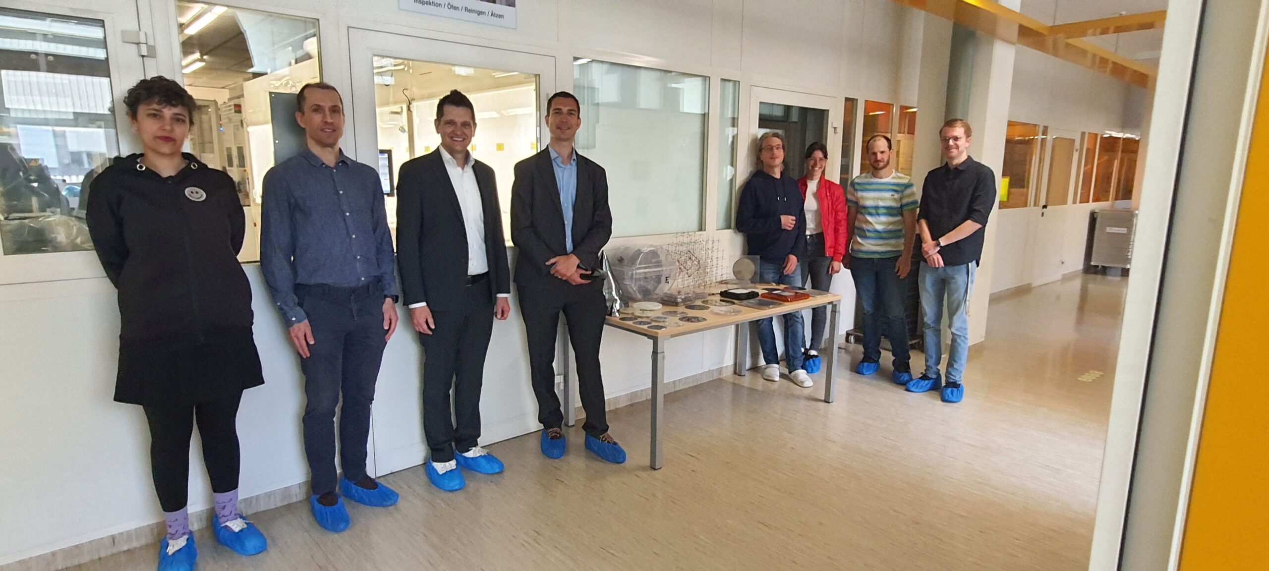 Prof. Dr. Brem (Executive Board IMS CHIPS & Vice-Rector for Transfer and International Affairs as well as Managing Director at ENI) and Prof. Dr. Nils Quack (Managing Director IMS CHIPS) are in the center of the picture, surrounded by employees of IMS CHIPS and colleagues from the Institute of Entrepreneurship and Innovation Science (ENI) at the University of Stuttgart. The group is located in the corridor along the windows to the clean room.